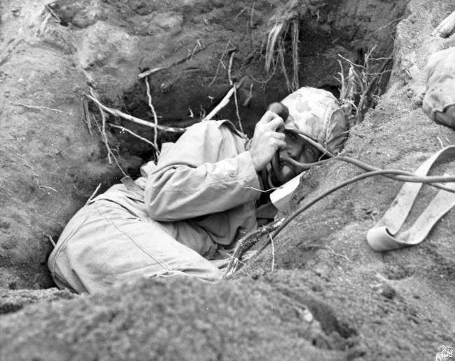 #25 This is a photo of a U.S. Marine communicator, burrowed in a shallow foxhole, calling for artillery support to silence enemy mortars in Iwo Jima, Japan, during World War II, 1945.