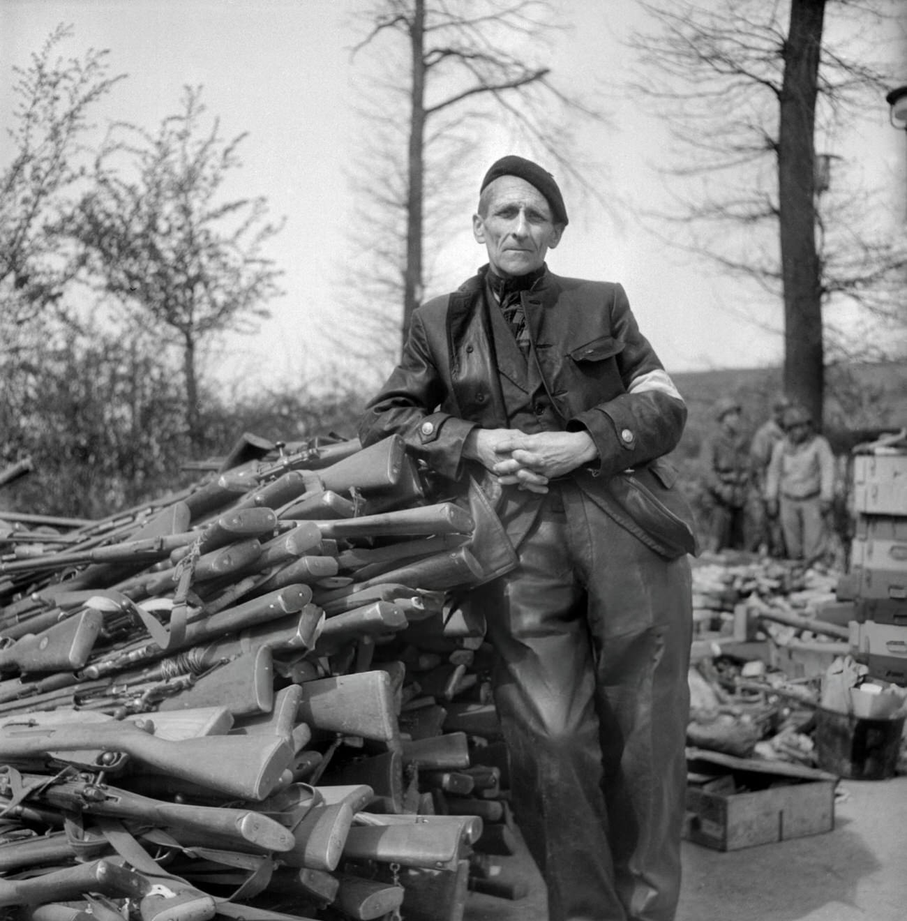 #37 An unidentified man poses in front of pile of weapons in the courtyard of the Buchenwald Nazi concentration camp, 1945.