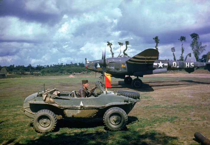 #17 A P-38 fighter plane sits in the background as the pilot arrives in a captured German vehicle, France, 1944