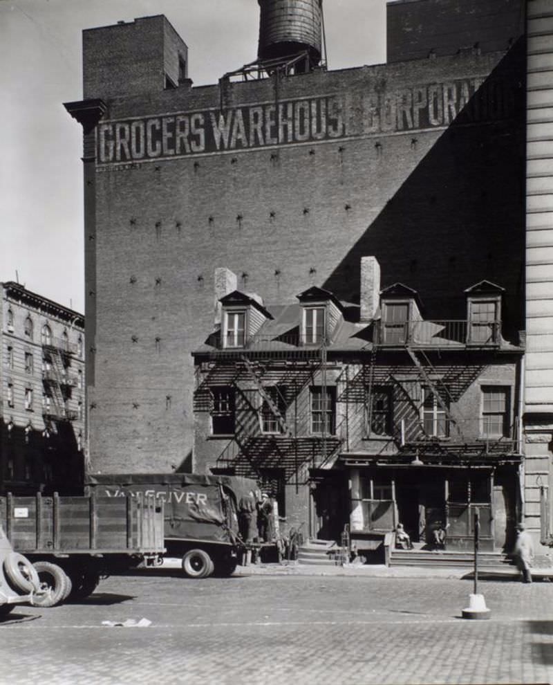 #1 Broome Street no. 512-514, Manhattan. Two row houses are dwarfed by the Grocers Warehouse Corporation building behind it, trucks in the foreground.