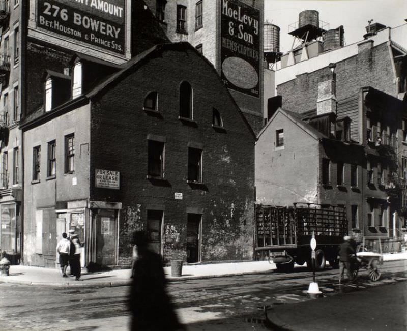 #15 Mulberry and Prince Streets, Manhattan. Building with gambrel roof, dormers on corner, small houses along street where man pushes cart, large buildings topped with water towers beyond.