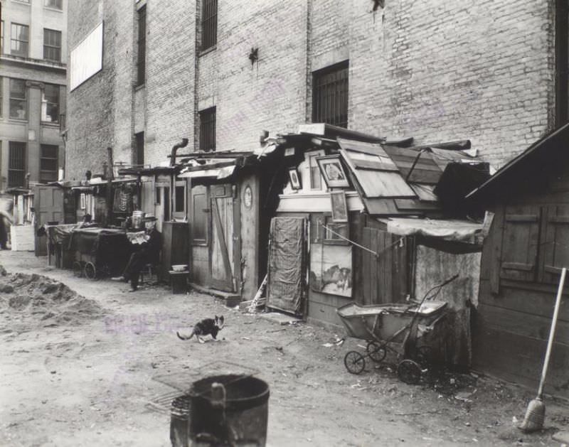 #70 Unemployed and huts, West Houston — Mercer St., Manhattan. Man sits reading in front of huts built of salvaged materials some decorated with pictures, cat in center, baby carriage, broom, right.