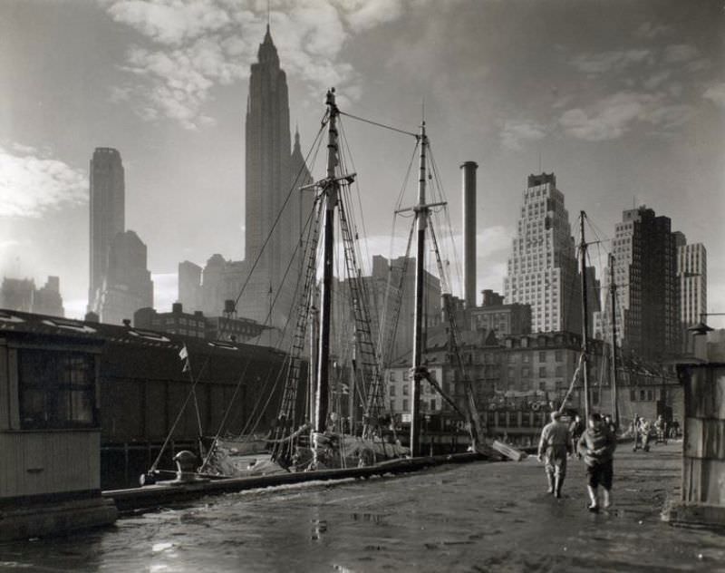 #21 Fulton Street Dock, Manhattan skyline, Manhattan. Men walk on pier where sailing vessels are moored, skyline beyond.