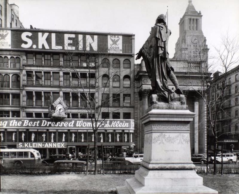 #29 Union Square, Manhattan. Looking up at statue of Lafayette from behind and left, S. Klein’s store, a bank, a hotel and the Consolidated Edison Building, beyond.