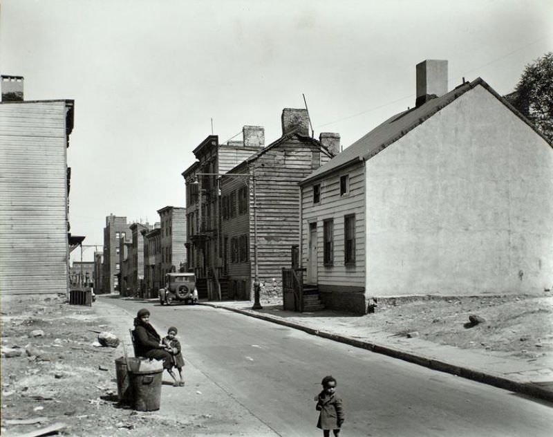 #60 Talman Street, between Jay and Bridge street, Brooklyn. African American woman sits at street edge with two children, empty lots on either side of street, old 2 and 3 story clapboard houses further up.