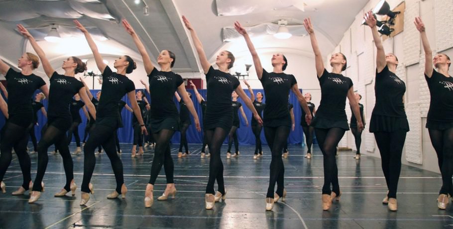 #76 A handful of Rockettes strike a balletic pose at rehearsal, at which they all wear standard-issue black outfits and footless tights. The floor’s tape marks complex blocking for the Christmas Spectacular routines.