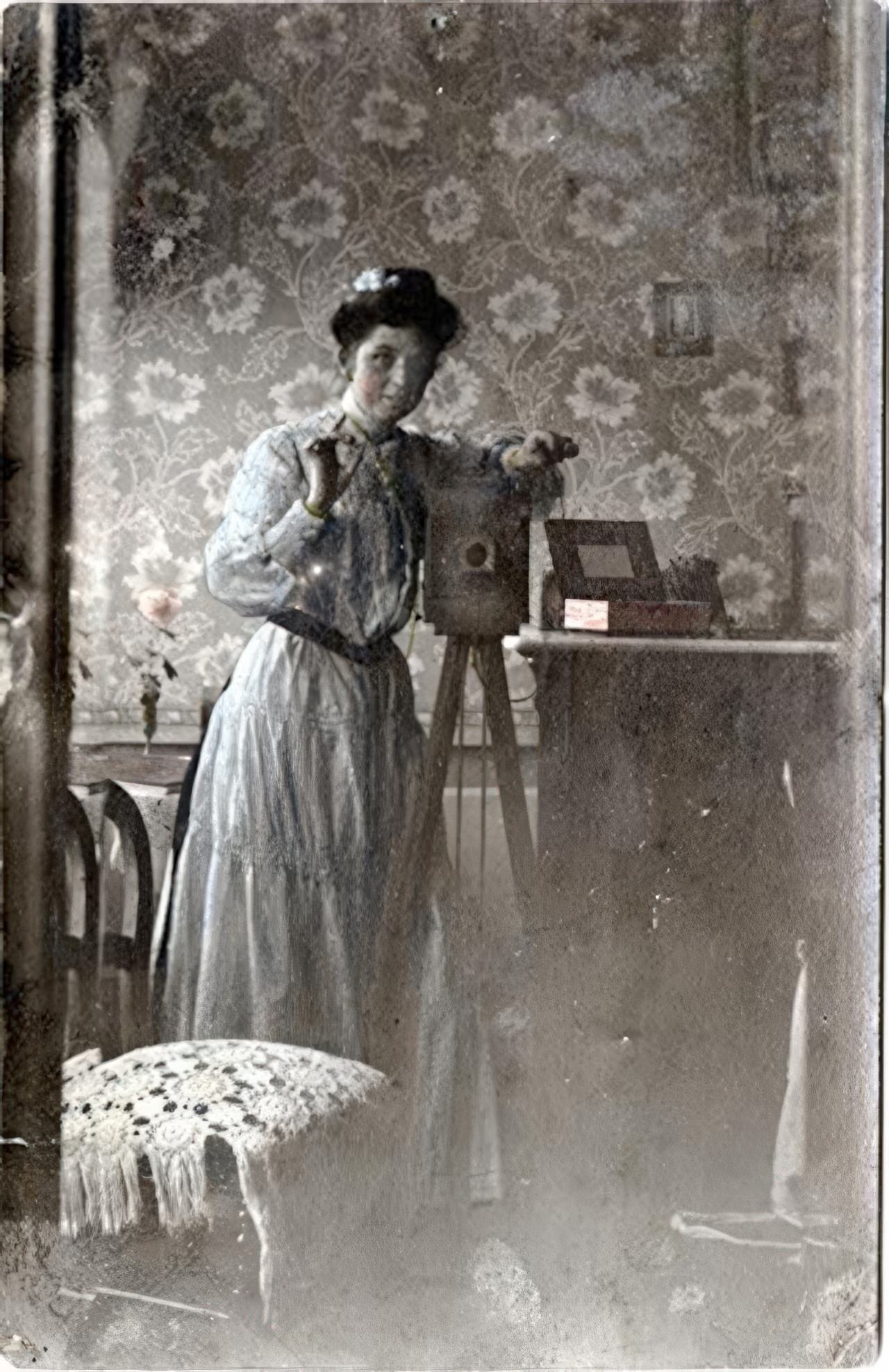 A young woman in a tinted blue dress and her camera are reflected in a large mirror. The left side shows a double image of a chair and the wall paper in the beveled edge of the mirror. This real photo postcard is marked “Rébublique Française” and “Carte Postale”.