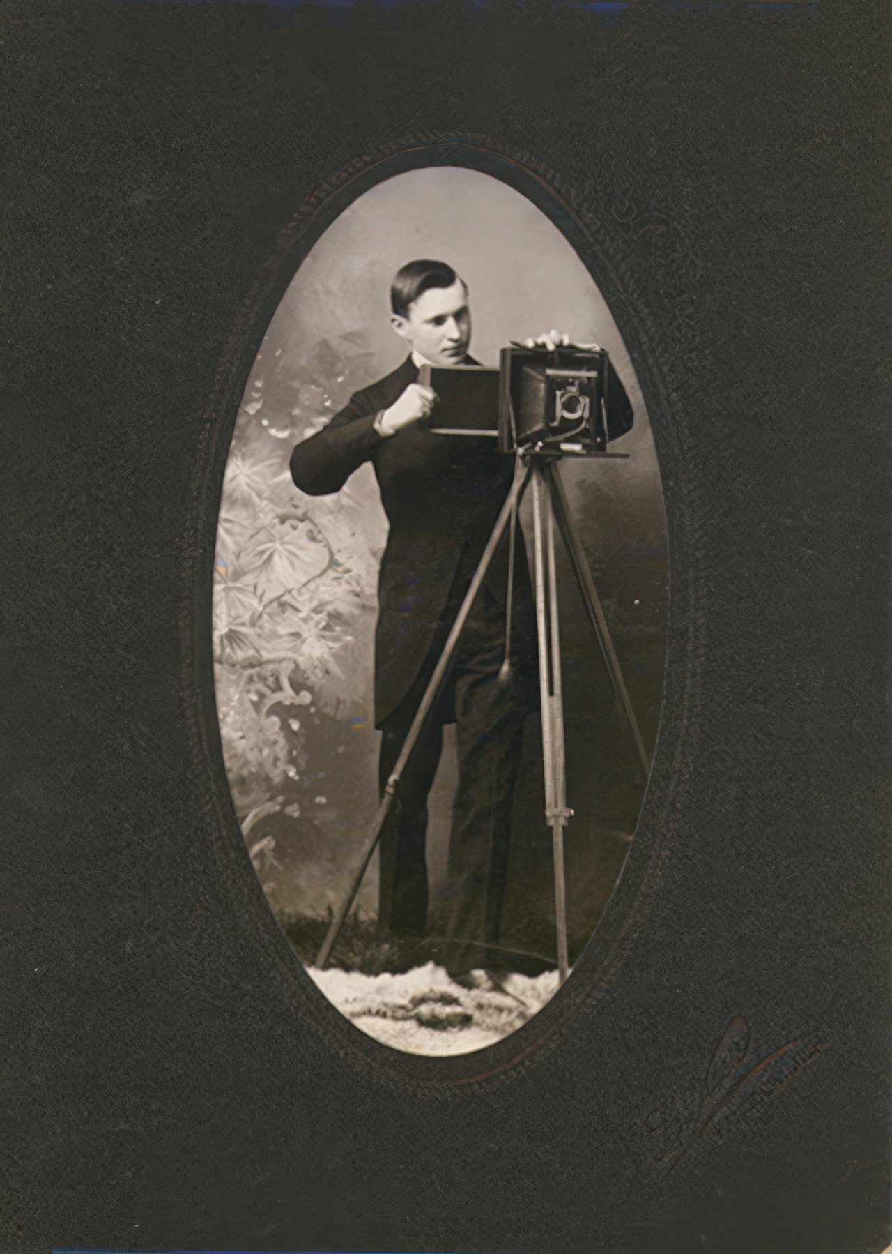 A well dressed young man inserts a film holder into a folding plate camera on a tripod in this oval cut photograph on a heavy black mat. The photographer’s name is embossed in hard to read script but the location, Hancock, Michigan is clear.