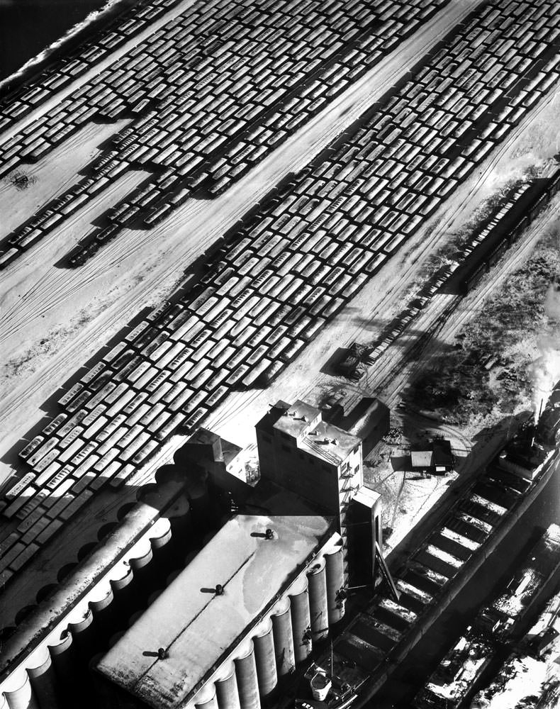#10 Grain elevator, operated by the Norris Grain Co. on the southeast side of Chicago, unloads corn from lake boat in a Calumet River slip (right foreground). In the freight yards (background) snow-covered gondola cars are loaded with coal.