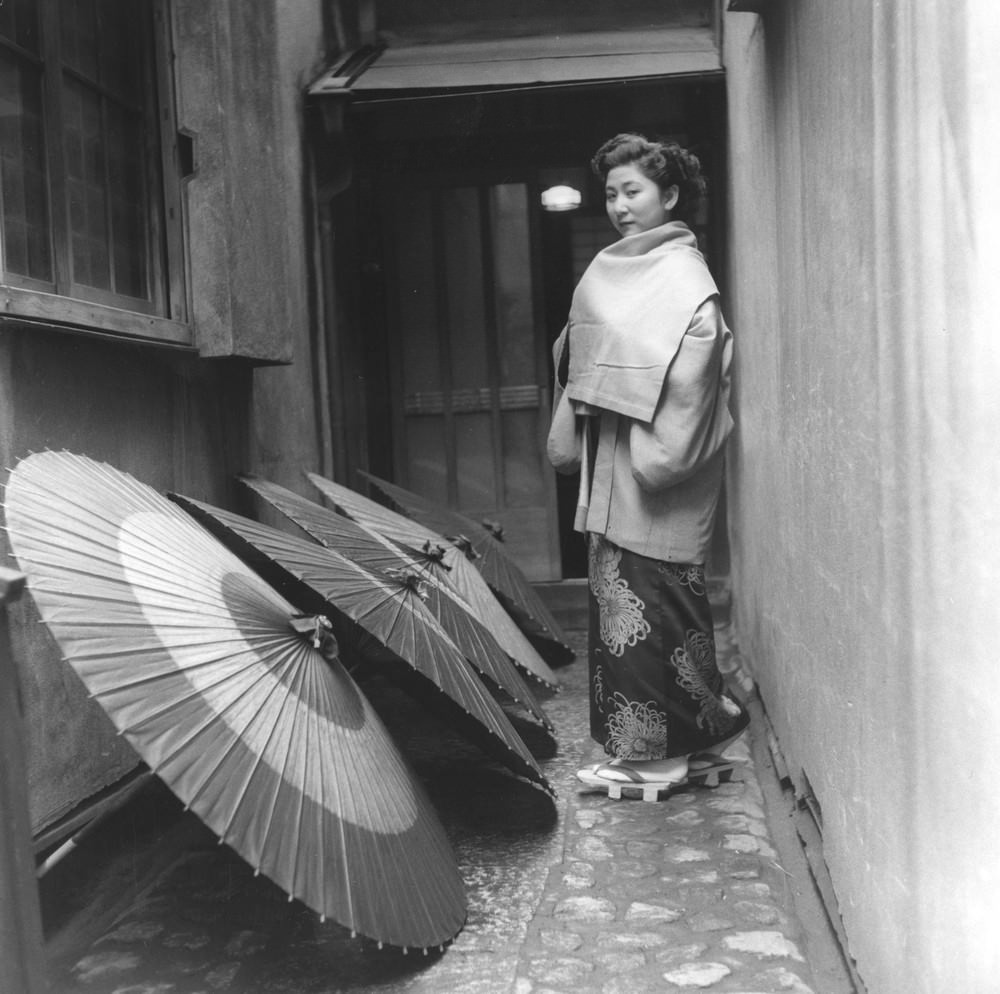#12 A Geisha passing drying umbrellas, 1950s.