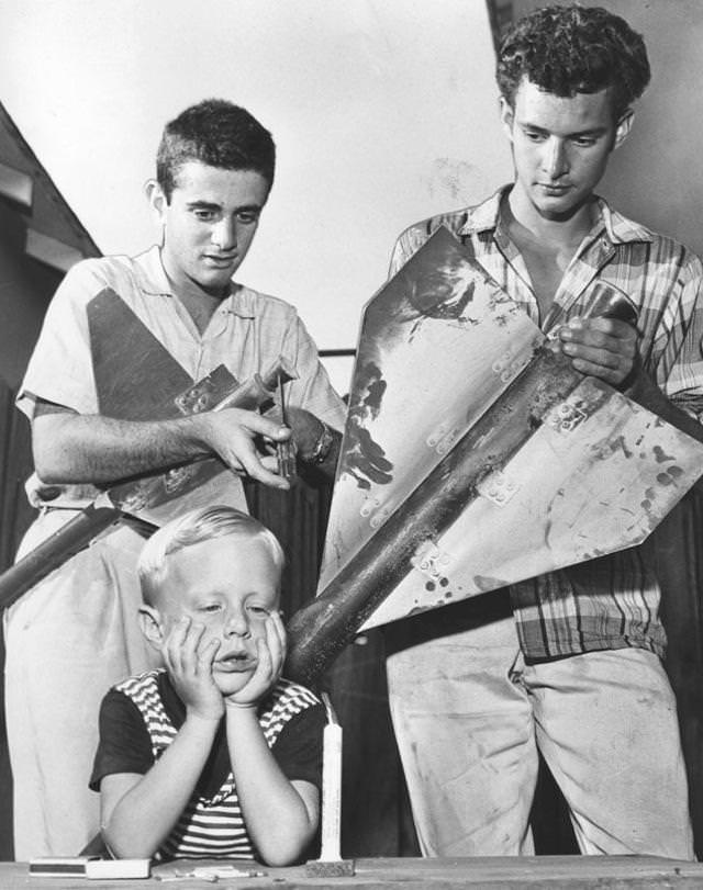 #11 Four-year-old Mark Malone of Burbank sticks to old-fashioned firecracker as his buddies, Myron Lieberman, 18, center, of Burbank, and Leston Newbill, 17, of Burbank, prepare to launch three-stage rocket they built for July Fourth celebration. 1959.