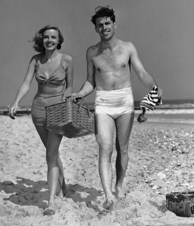 #14 Circa 1950s: Couple walking with picnic basket on beach.