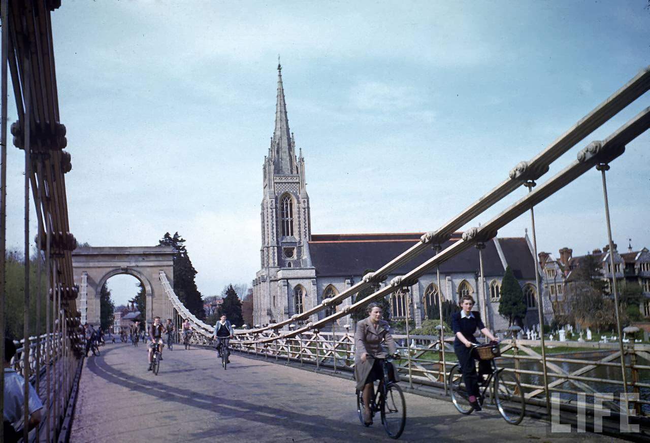 Cyclists crossing suspension bridge past unident. church during WWII.