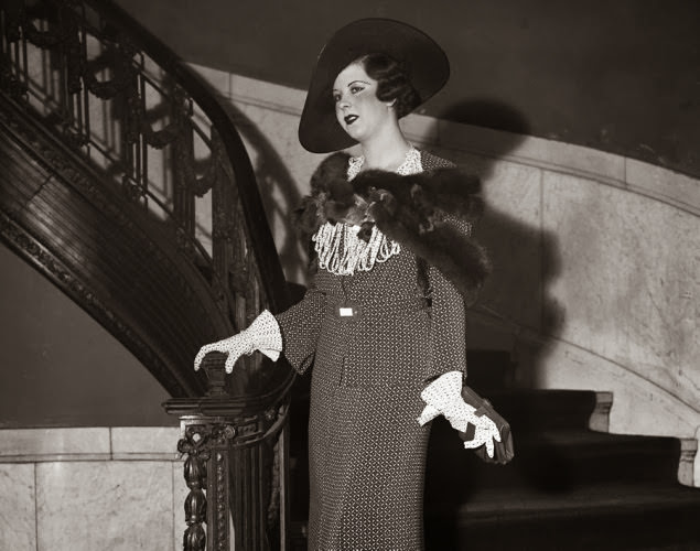 #9 Model Jean Early poses on the staircase at a 1934 summer fashion show while completely decked out in an elegant suit with a fur cape and hat.