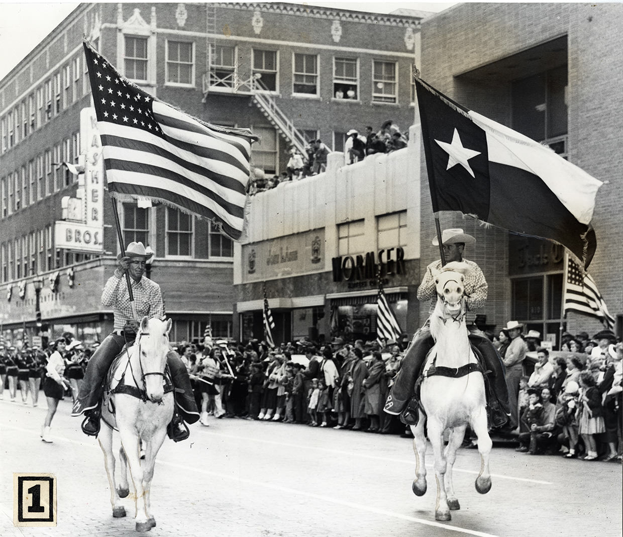 #67 Two men on white horses in Stock Show parade carrying United States flag and Texas flag, downtown Fort Worth, Texas, 1953.