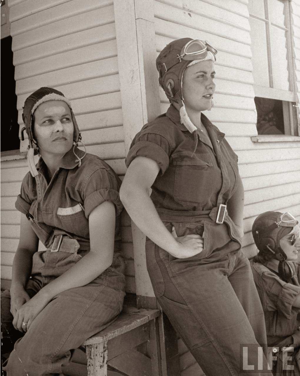 #1 Rebecca Edwards, 22, w. cream on her sunburned face & Lorena Daly, both pilot trainees in the Women’s Flying Training Detachment, sporting goggled leather helmets & G.I. coveralls as they lean against primary hangar fr. which they fly, at Avenger Field.