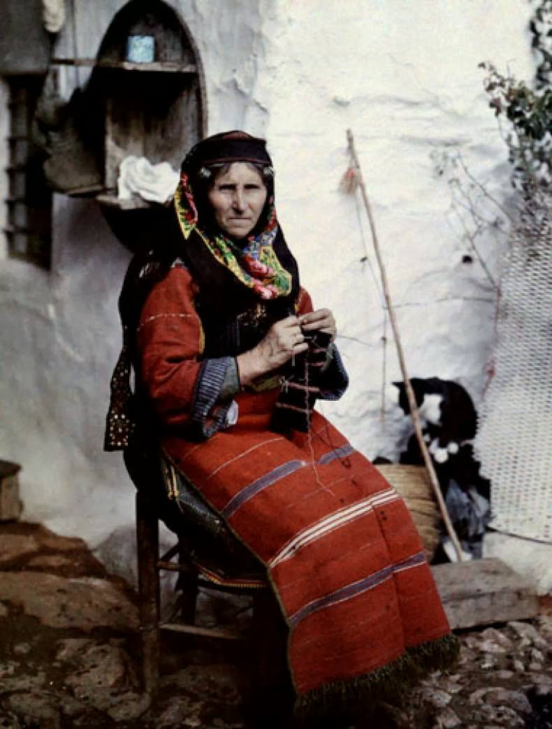 A woman in traditional Greek dress knits sitting outside her home, Aivali.