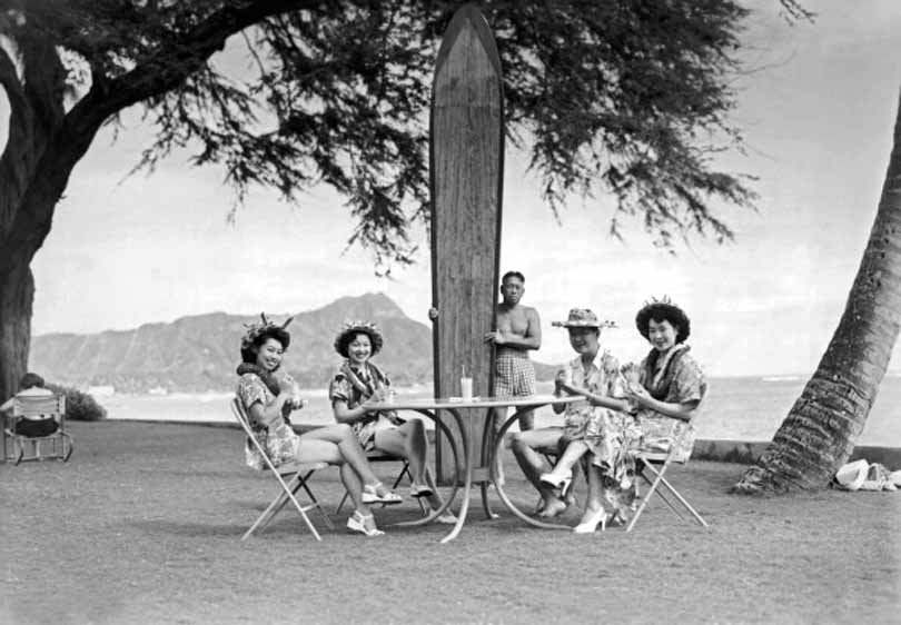 #80 Four Japanese tourists pose at the Halekulani Hotel on Waikiki Beach with a native Hawaiian and his long surfboard, 1930.
