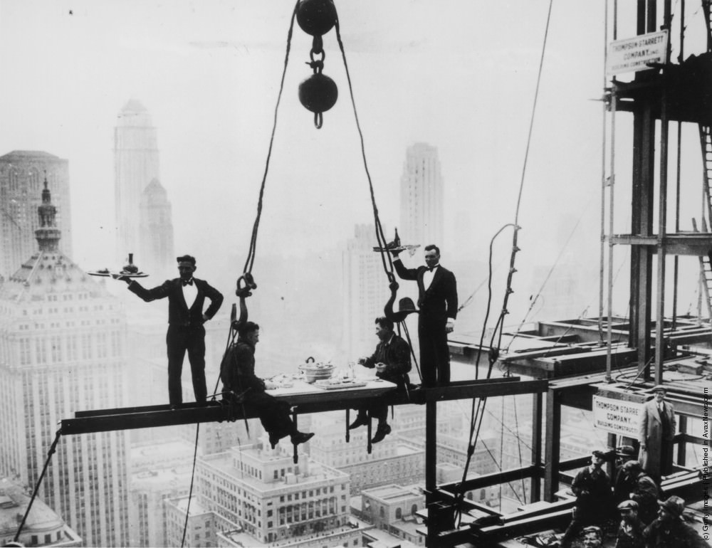 Two waiters serve two steel workers lunch on a girder high above New York City, the Waldorf-Astoria Hotel under construction, 1930.