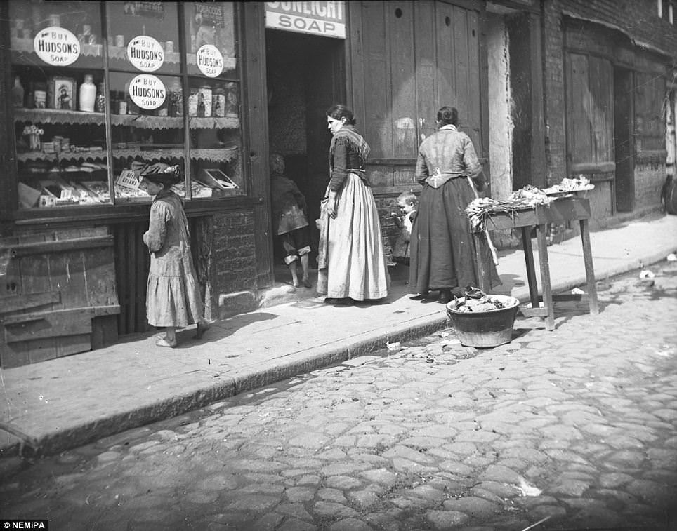 #7 A young girl examines the window display of a city shop selling fresh veg, sunlight soap and sweets.