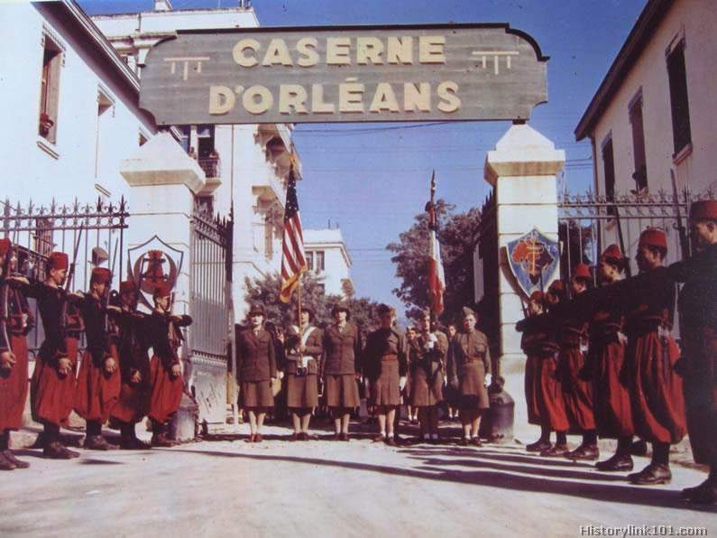 The Color Guard of the Women’s Army Corps in North Africa is seen coming through the gate of the Caserne D’Orleans, a famous Mediterranean fortress, while the guard of honor of the First Zouave Regiment stands at attention.