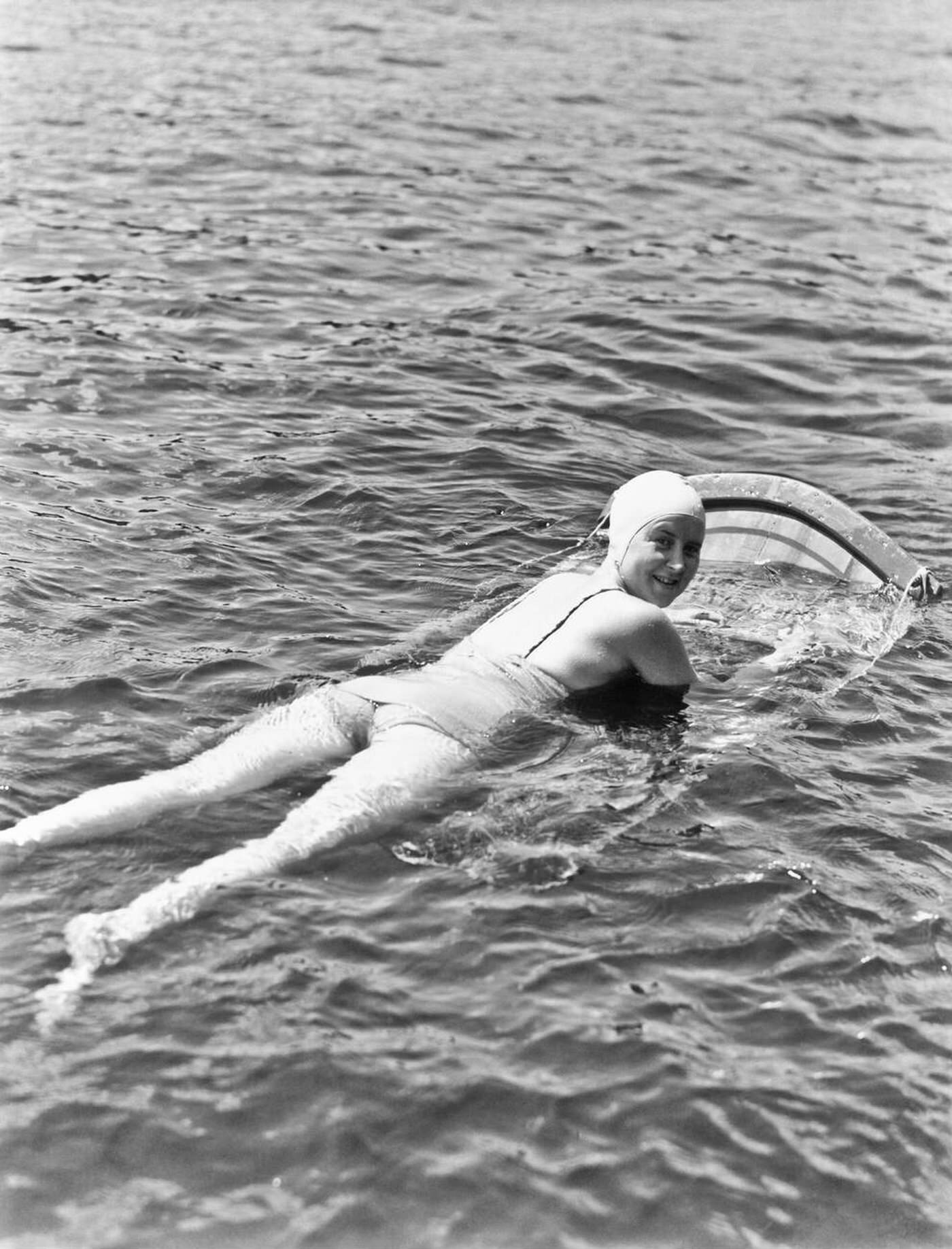 #15 Woman wearing a bathing cap and one-piece suit lying on an aqua ski board looking at the camera over her shoulder at Muskoka Lake, Canada, 1935.